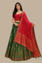 A woman models a traditional Indian ethnic wear set consisting of a green and red Banarasi silk lehenga choli with a matching red dupatta, set against a background of a clothing store with folded garments.
