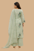 A woman stands with her back to the camera wearing a light sage green three-piece traditional outfit with floral embroidery, consisting of a kurta, pants, and a matching dupatta, inside a fabric store.