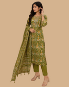 A woman models a green printed salwar kameez suit with matching pants and a dupatta inside a clothing store. The suit features a detailed paisley and floral pattern in shades of green, yellow, and orange.